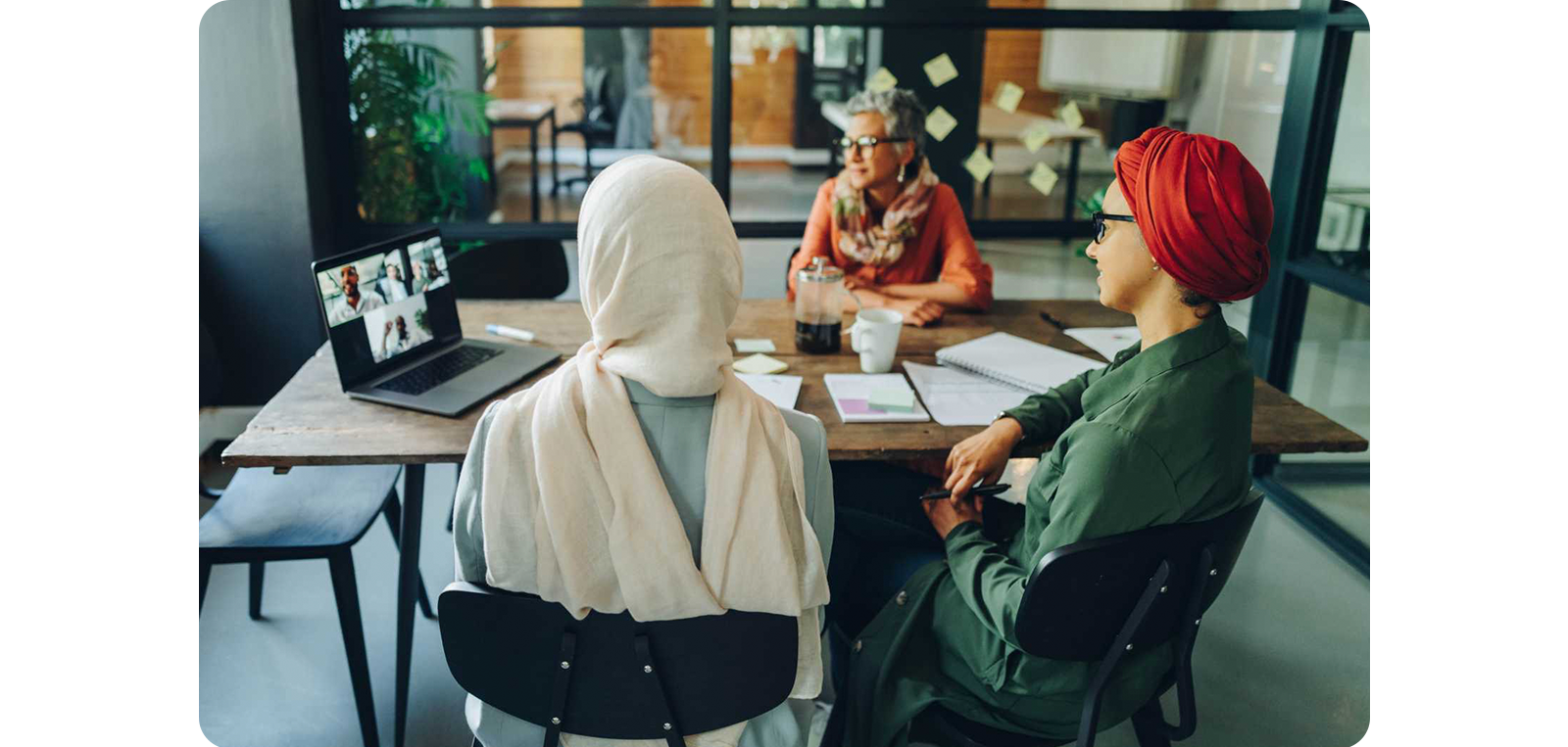 Three individuals are participating in a diverse and inclusive group meeting. They are seen from behind, looking towards a third participant on a laptop screen, illustrating a hybrid meeting setup that ensures equitable participation opportunities for all members, regardless of their physical location. The setting conveys a sense of digital connectedness and the facilitation of inclusive dialogue.