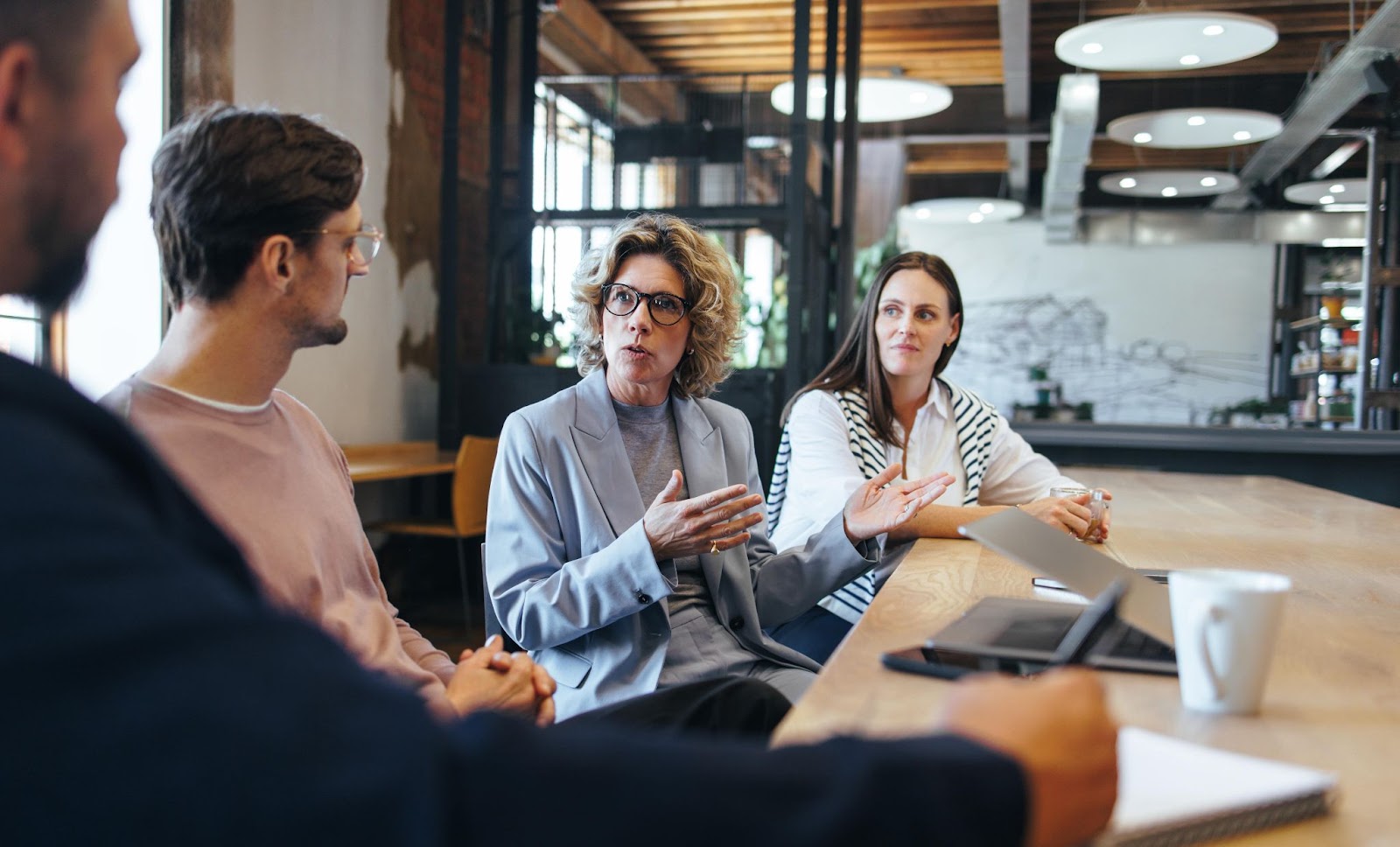 A diverse group of professionals is engaged in a discussion around a wooden table in a modern office space. A woman in a gray blazer gestures as she explains a point, her colleagues listening attentively, illustrating a collaborative learning environment where knowledge and ideas are being shared to impact organizational culture and behaviors.