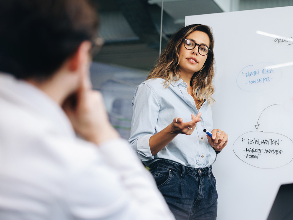 A professional trainer in a light blue shirt and glasses stands confidently in front of a whiteboard, gesturing towards a diagram.